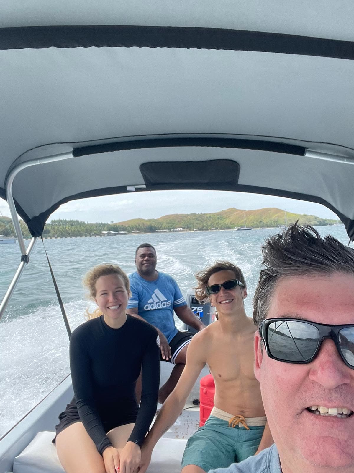 Group selfie on the boat cruising past a green Fijian island, wake trailing behind